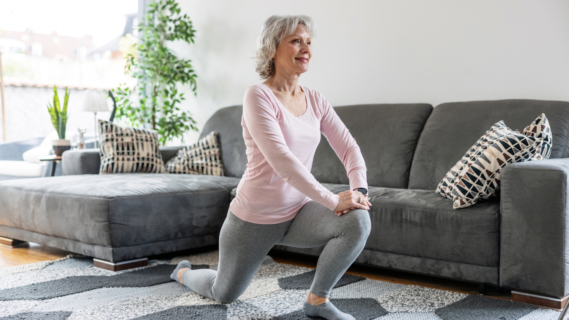 woman wearing a light pink top and grey leggings performing a deep forward lunge in a living room. there's a grey sofa behind her and grey patterned rug beneath her. 